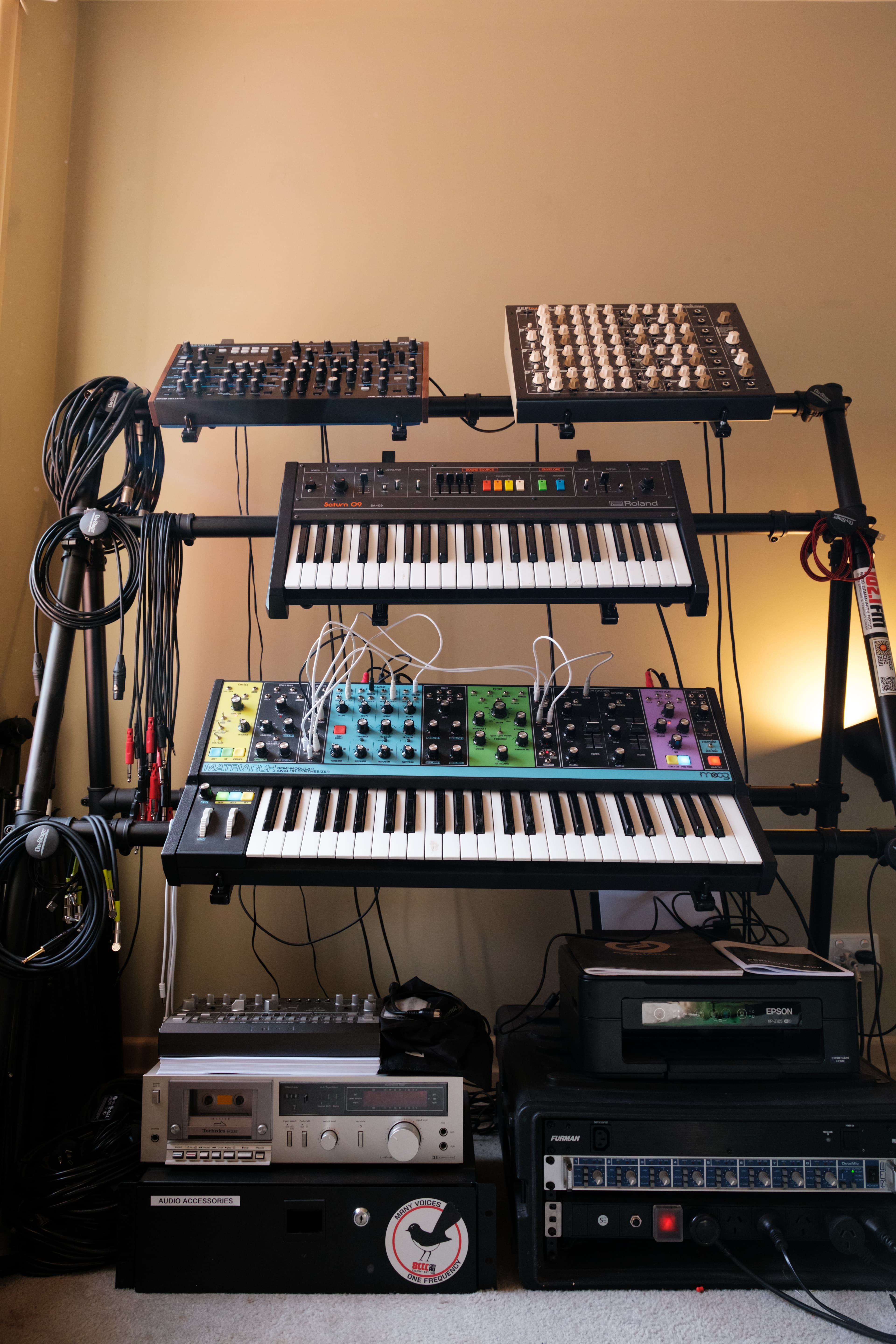 Rack of analog synthesizers, a cassette deck, and a mixer against a studio wall
